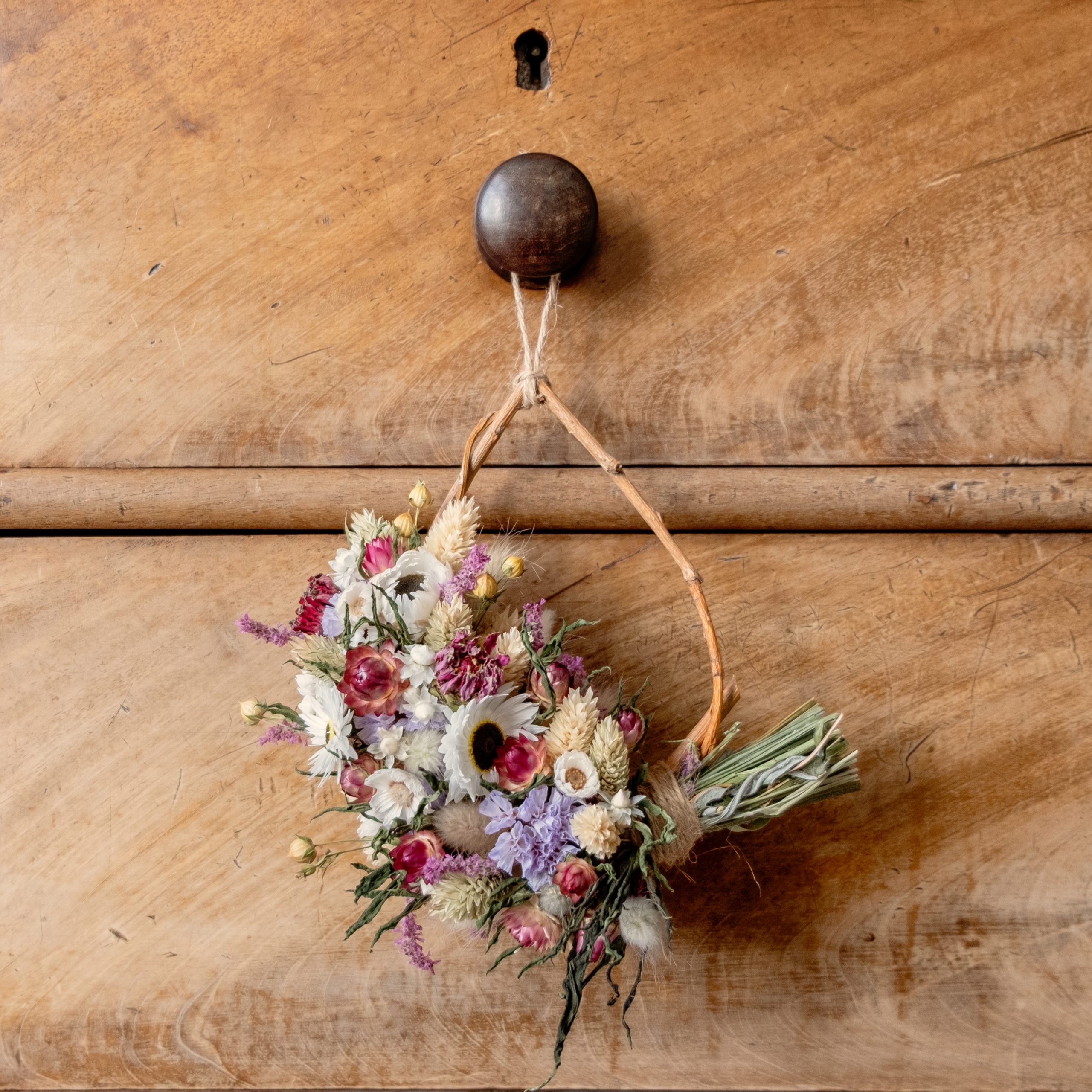 Mini tear drop shaped willow wreath. The wreath is half covered in dried flowers and hung on a wooden drawer knob