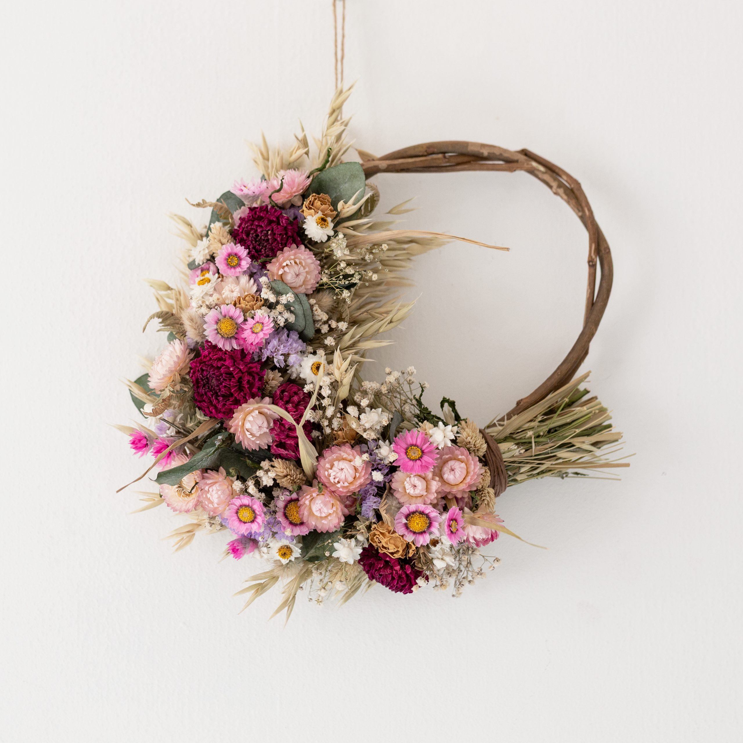 dried flower wreath made from pink and lilac flowers, with white flowers and eucalyptus dotted around. the wreath is half covered in flowers with a natural willow hoop. its hung a white wall