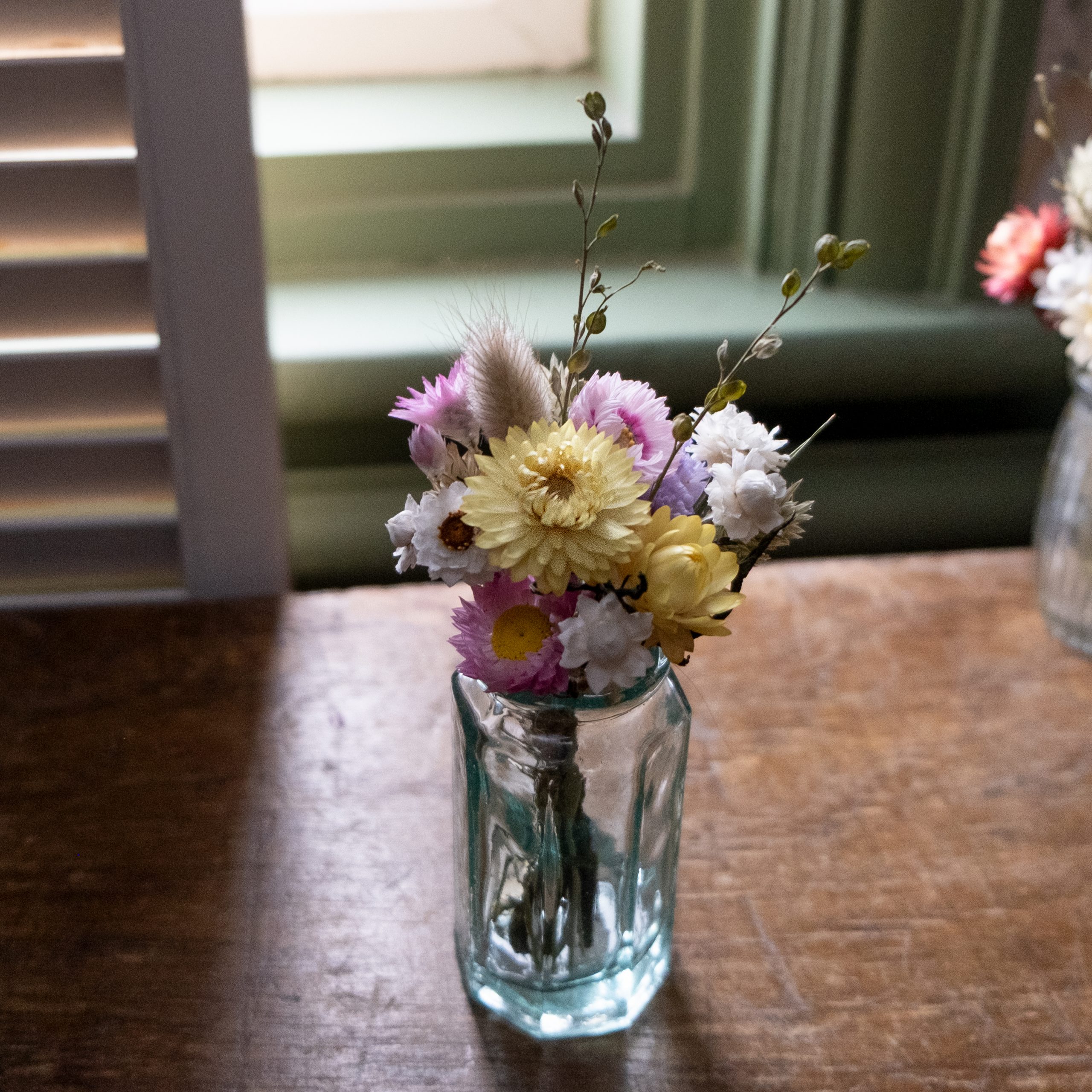 Clear glass bud vase with a mini yellow pink and white posy. on a wooden surface with a window in the background.